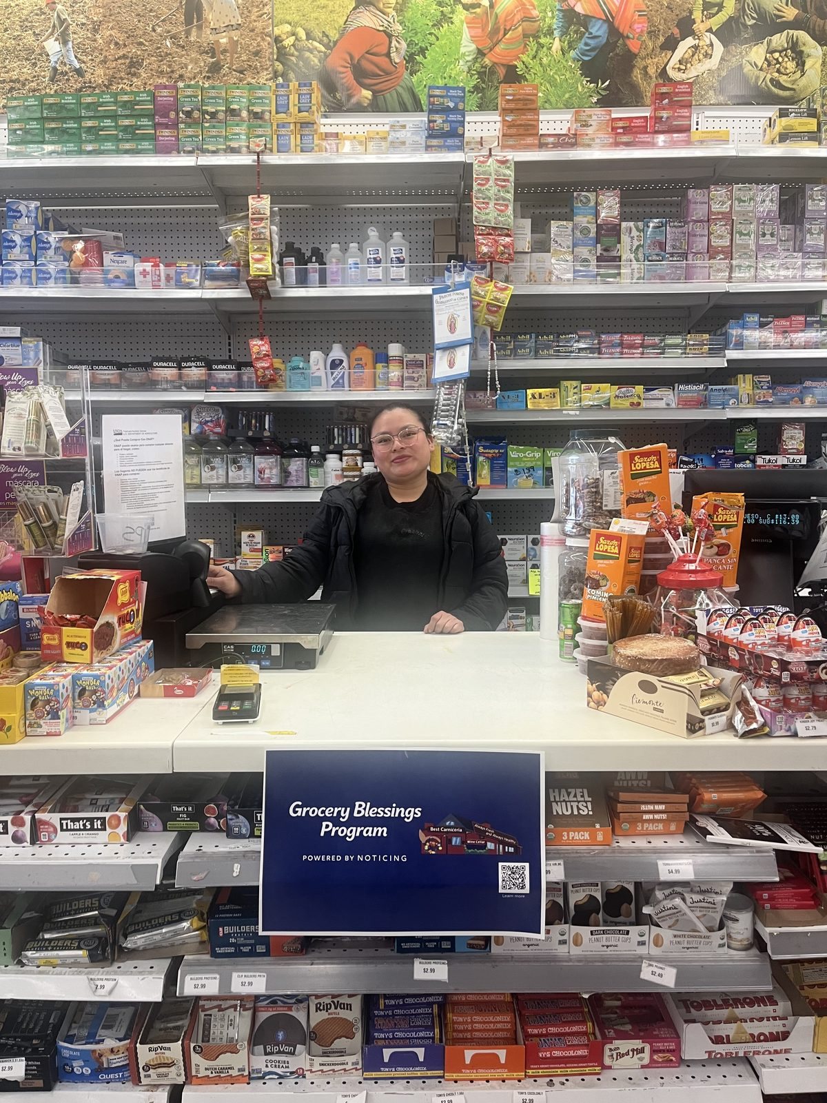 A cashier at Casa Guadalupe behind the counter, with a Grocery Blessings Program sign on the register.